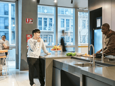 Two men in a kitchen, one cooking, the other eating fruit, and two women seated nearby.