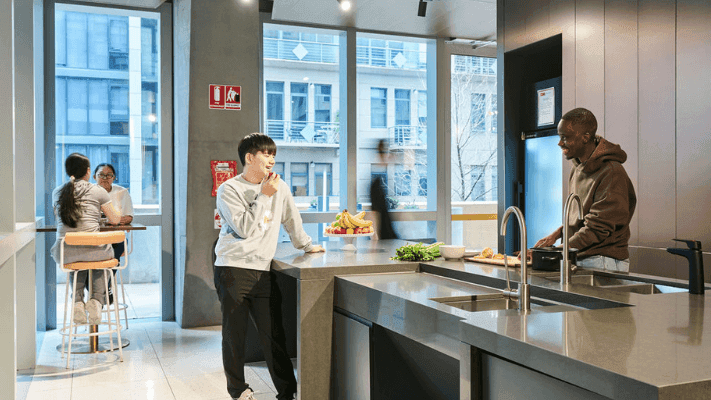 Two men in a kitchen, one cooking, the other eating fruit, and two women seated nearby.
