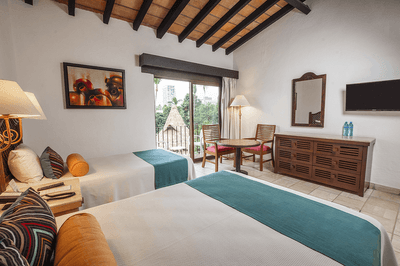 Two beds with colorful pillows, nightstands, dresser, mirror, TV, and balcony with chairs and table.