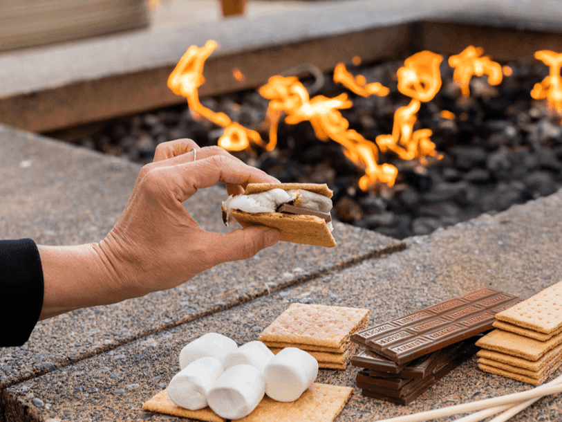 freshly made s'more with melted marshmallow and chocolate between two graham crackers in front of a fire pit