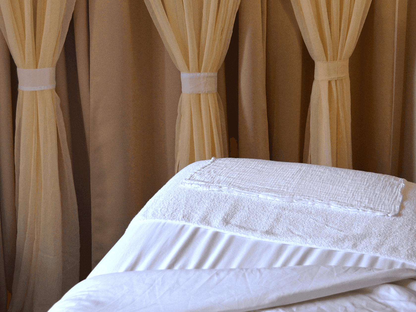 White towel and sheet on a bed in front of beige curtains.