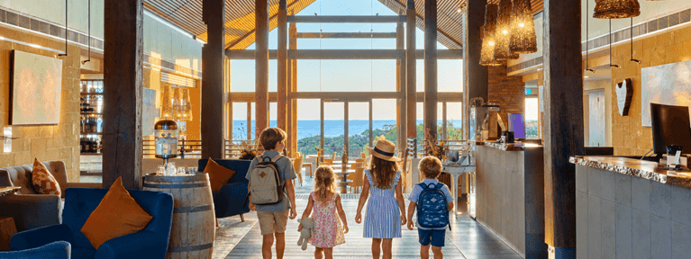 Family with children walking through a bright hotel lobby with wooden beams and ocean views during Spring School Holidays