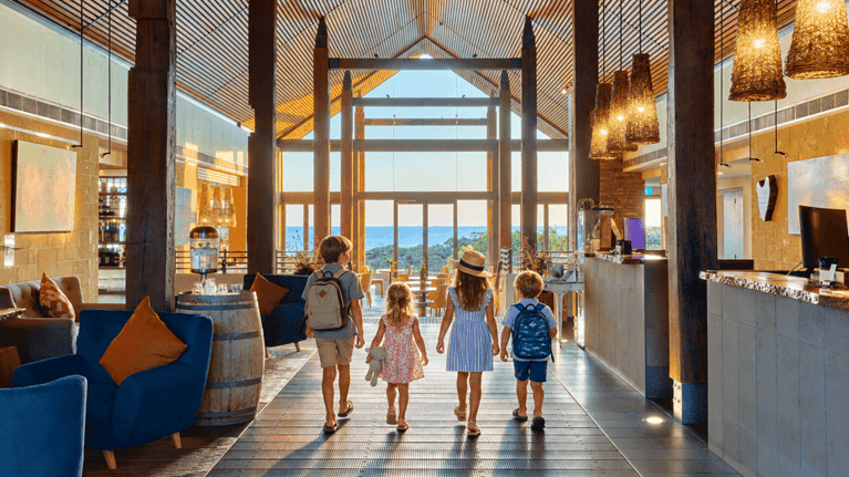 Family with children walking through a bright hotel lobby with wooden beams and ocean views during Spring School Holidays