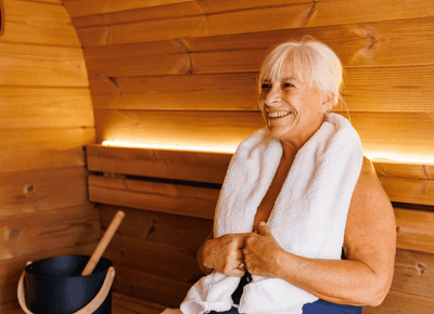 Smiling woman resting with a soft white towel around her neck beside a water ladle inside the sauna at Rodd Moncton