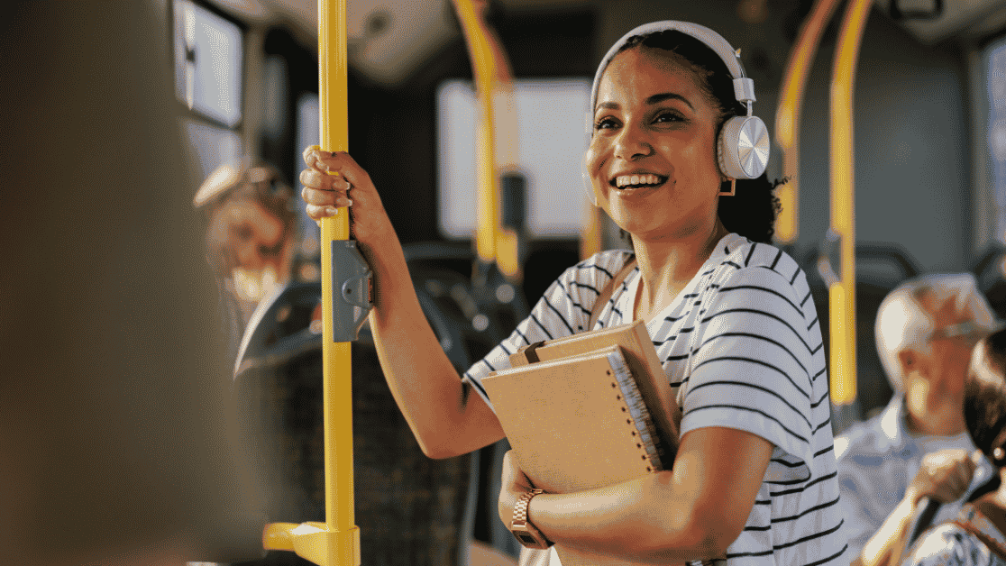 Smiling student with headphones holding books while standing on a bus handrail.