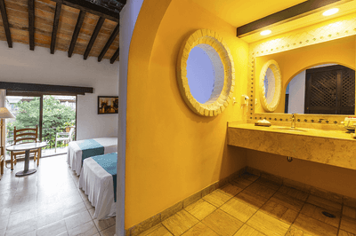 A yellow-themed room with a bathroom featuring round mirrors, and a bedroom with two beds and a balcony.