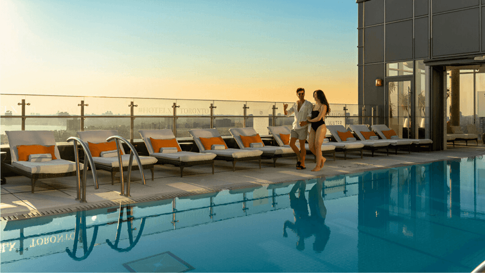 Couple walking together beside rooftop pool with white sun loungers facing scenic glass balcony view at Hotel X Toronto
