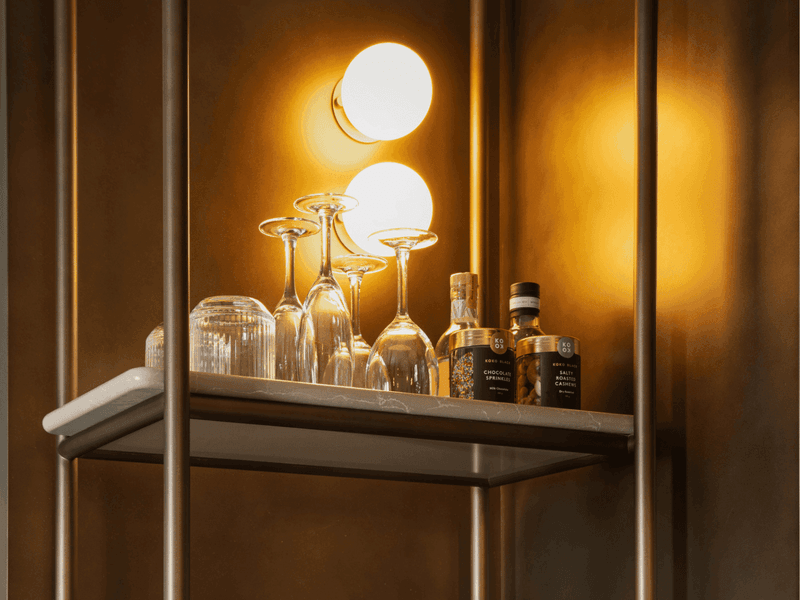 Hotel minibar shelf with wine glasses, shot glasses and premium liquor bottles under warm lighting