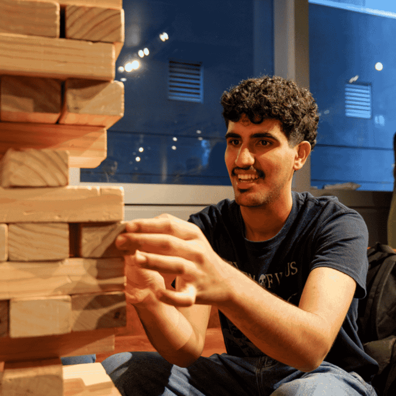 Young man playing giant Jenga in a modern communal lounge with blue walls and large windows at UniLodge