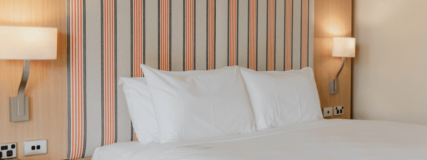 A white hotel bed with two pillows, a nightstand with a lamp and telephone, and striped wall.