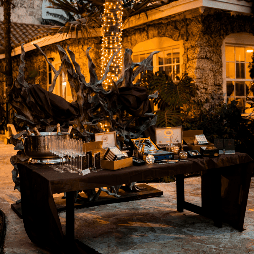 Evening outdoor event setup with champagne glasses, cigars, and decorative items on dark tables beneath illuminated trees.