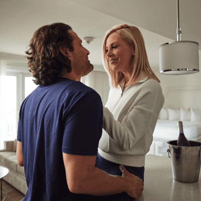 brunette man and blonde women in an oceanfront suite getting cozy with a bottle of champagne