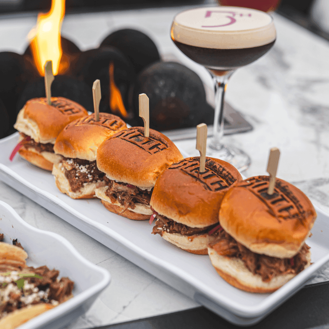 A plate of sliders and a drink on a table with a fire pit.