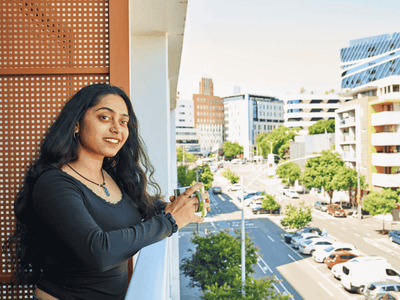 Woman on balcony at UniLodge Royal Melbourne overlooking street with cars and buildings.