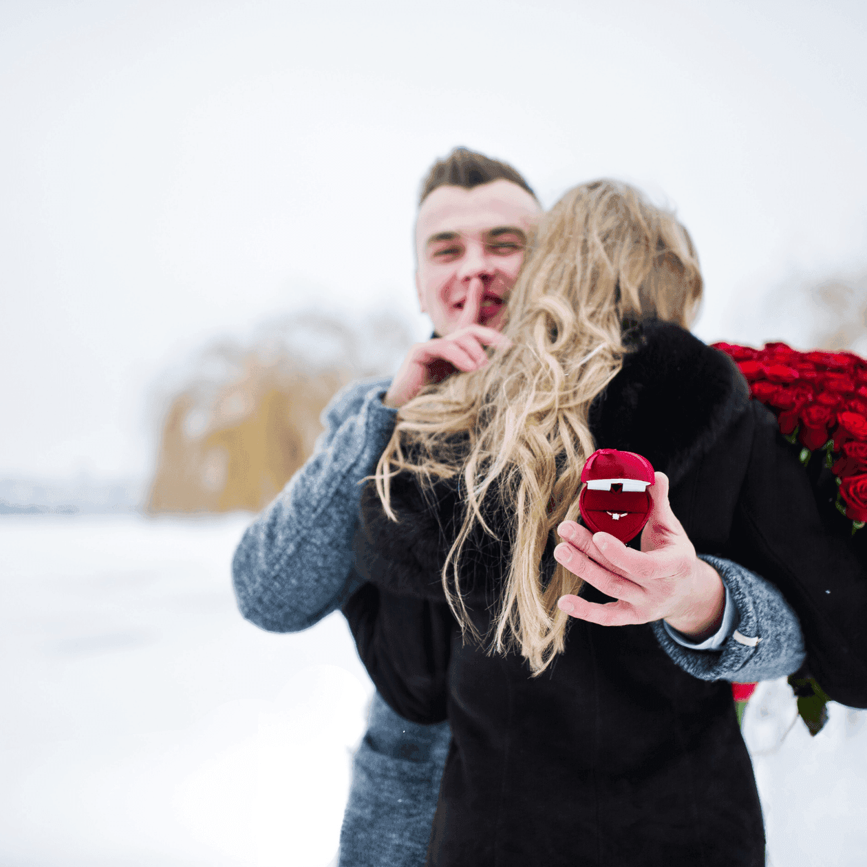 Couple in snowy field holding engagement ring and red roses.