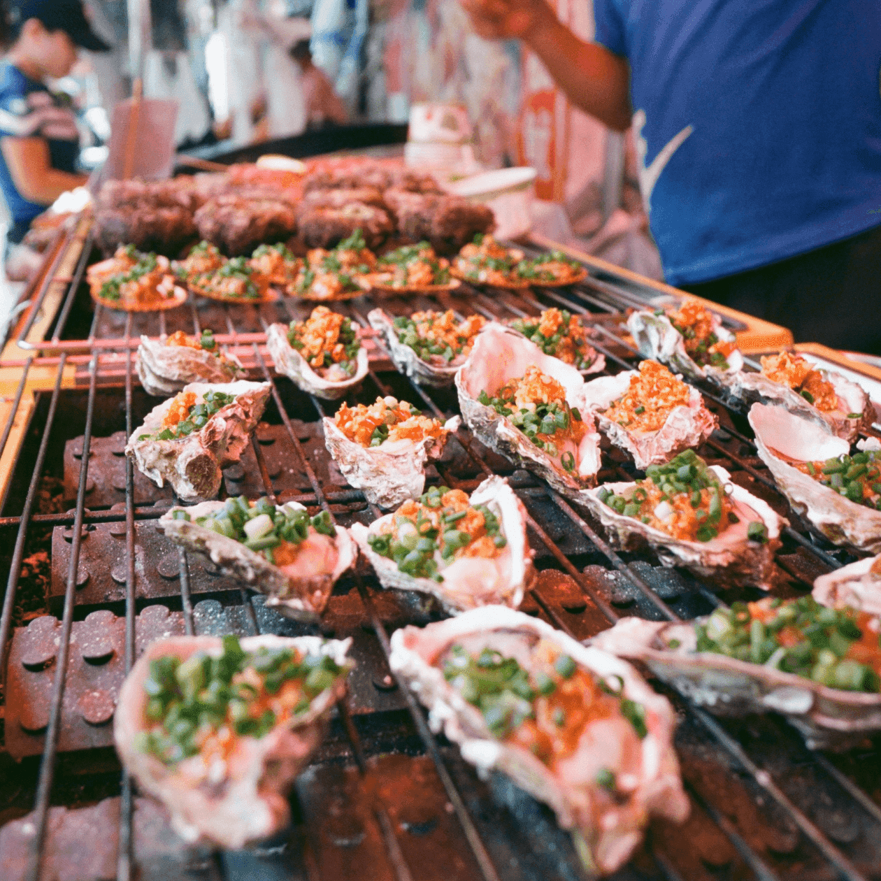A street food vendor displaying seafood skewers and oysters on a grill, with customers in the background.