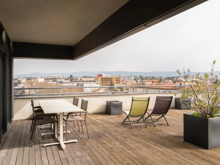 Outdoor wooden deck with dining table and lounge chairs, overlooking cityscape at Appart’Hôtel Hévéa, Valence.