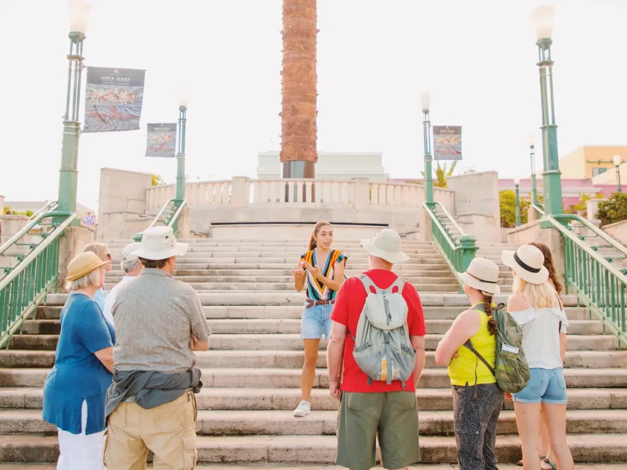 Grupo de turistas con sombreros y mochilas observando una torre de ladrillo histórica desde una plaza con escalinata.