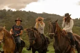 Three riders atop brown horses wearing hats, with forested hills and cloudy skies surrounding them near Unika Hotels