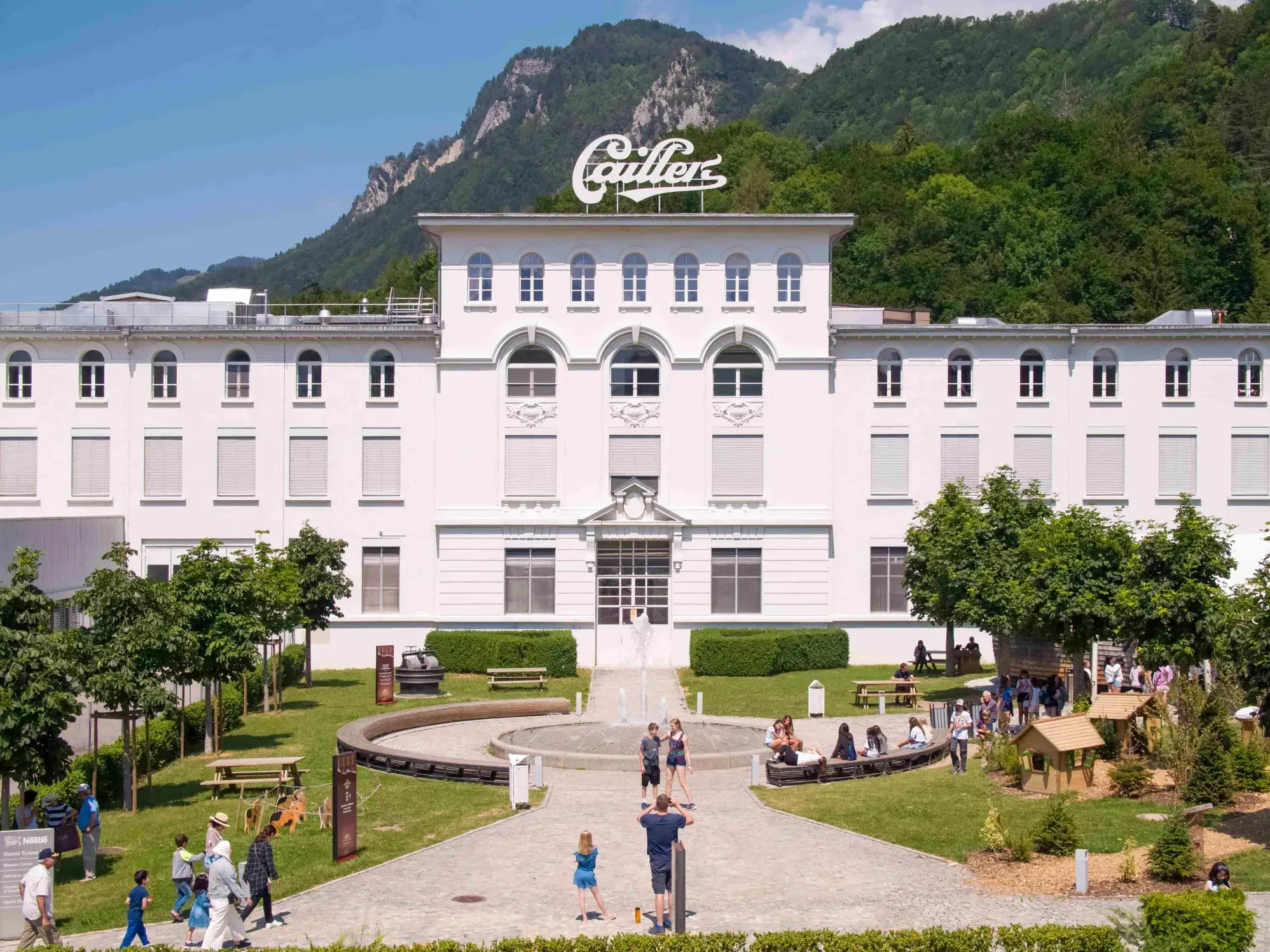 Exterior of Maison Cailler surrounded by lush greenery, and visitors near Hotel des Innovations