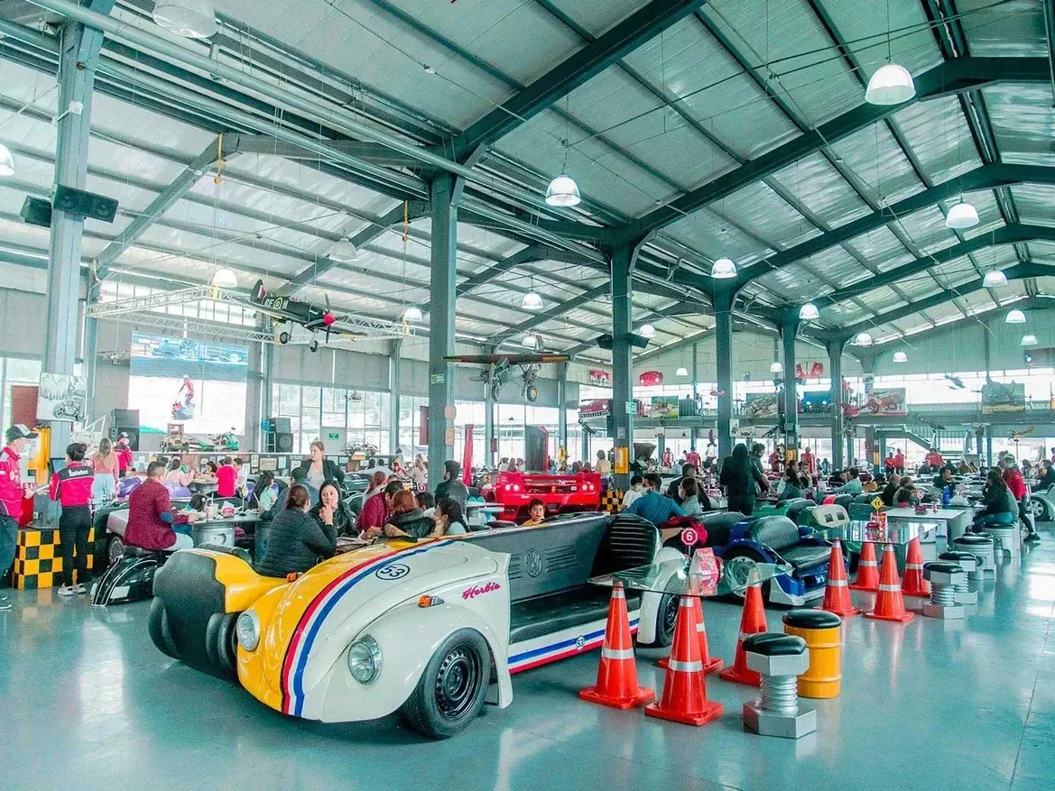 Amplio espacio interior de un restaurante o centro de entretenimiento con mesas y personas, destacando cabinas únicas con temática de coches clásicos y un techo alto