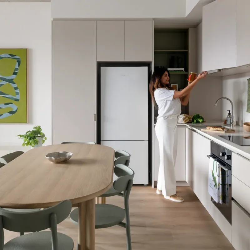 Woman reaching for a bowl in a modern kitchen with white cabinets and wooden table.