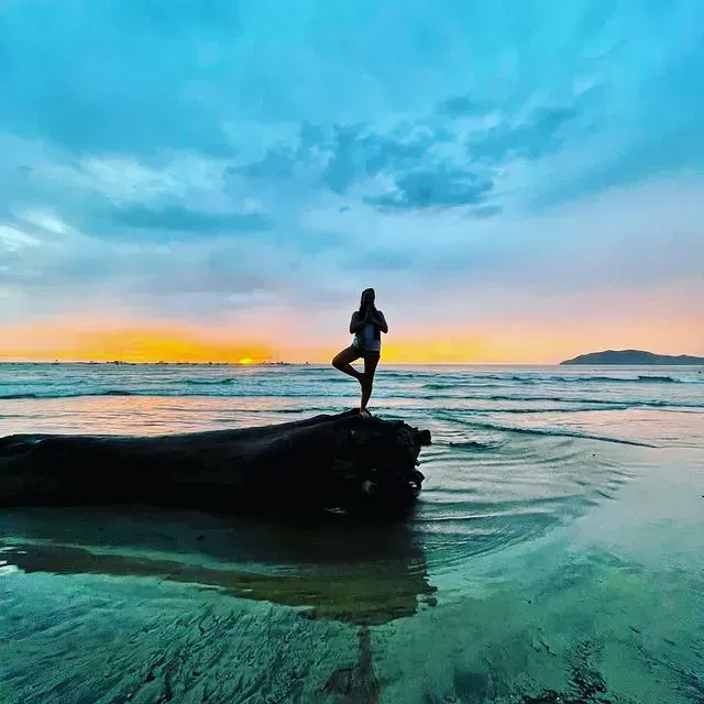 Person in tree pose on driftwood at beach during sunrise with turquoise water and colorful sky at El Blok Hotel in Vieques.