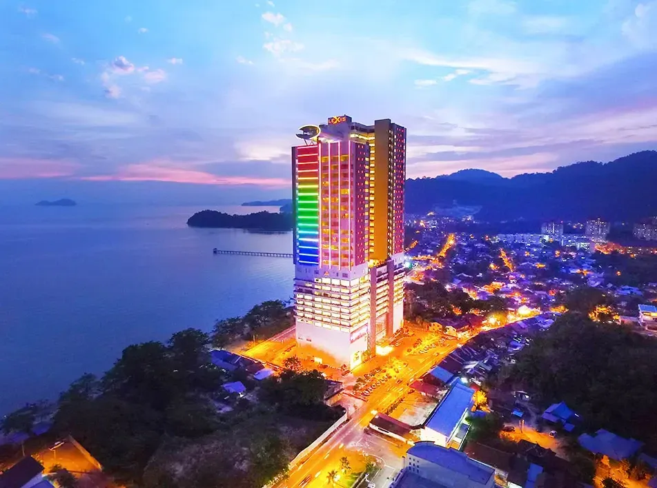 Colourfully lit Lexis Suites Penang hotel tower with lights by the sea at dusk.