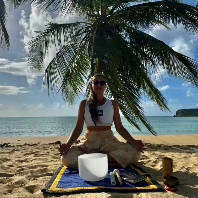 Woman meditating on beach with singing bowls under palm trees during Yin & Restore Sunset Sound Bath session at El Blok Hotel.