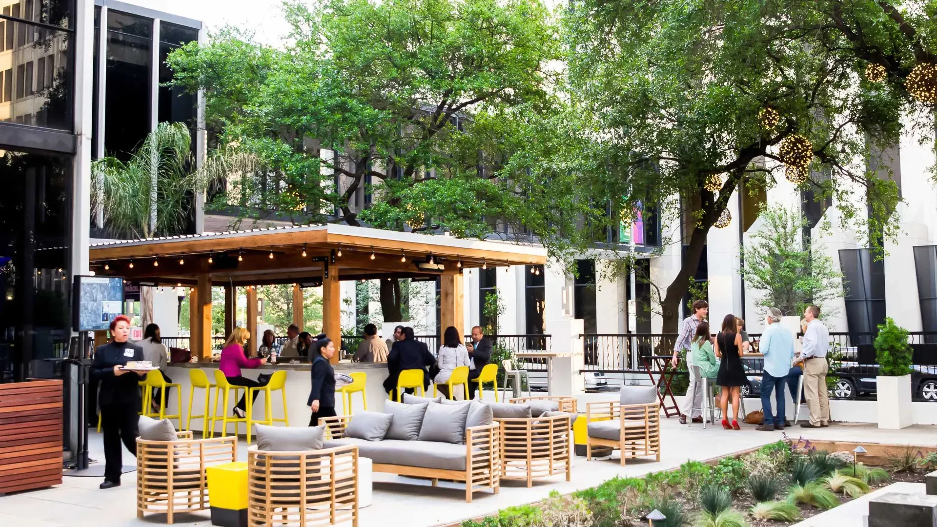A group of people socializing in an outdoor bar area with a pavilion, surrounded by trees and modern buildings.