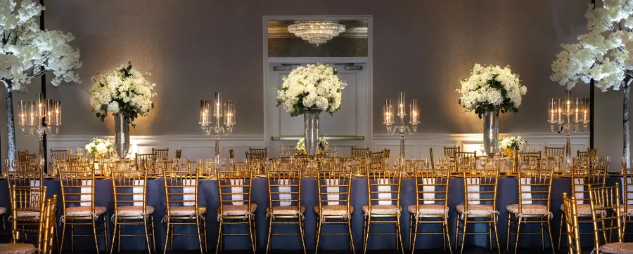 Long dining table by gold chairs under white floral towers in Turtle Creek Ballroom at Warwick Melrose Dallas