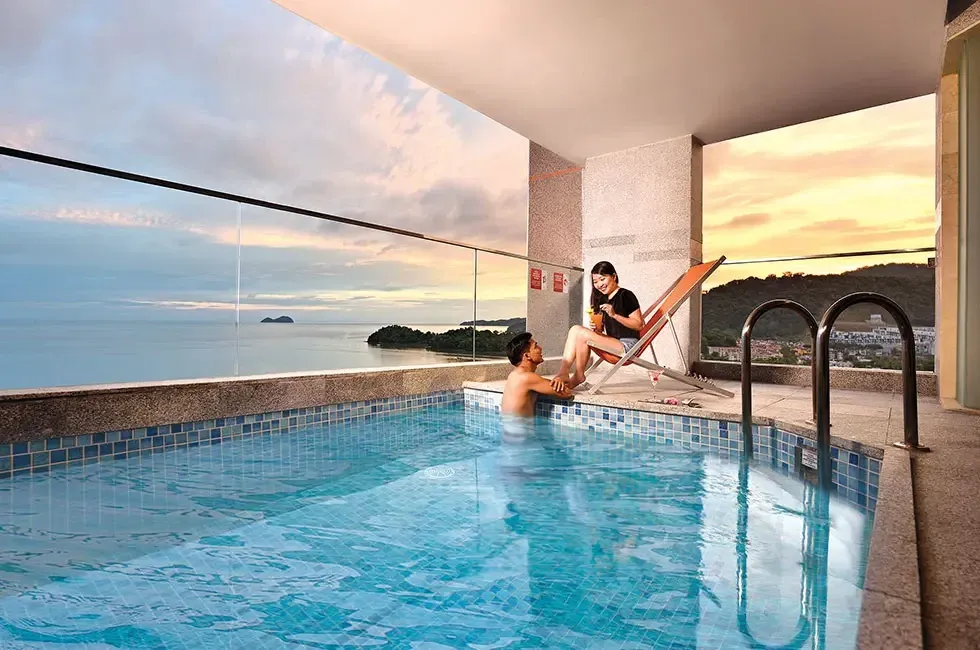 Couple relaxing by a rooftop pool overlooking the ocean at sunset at the Lexis Suites Penang Hotel.