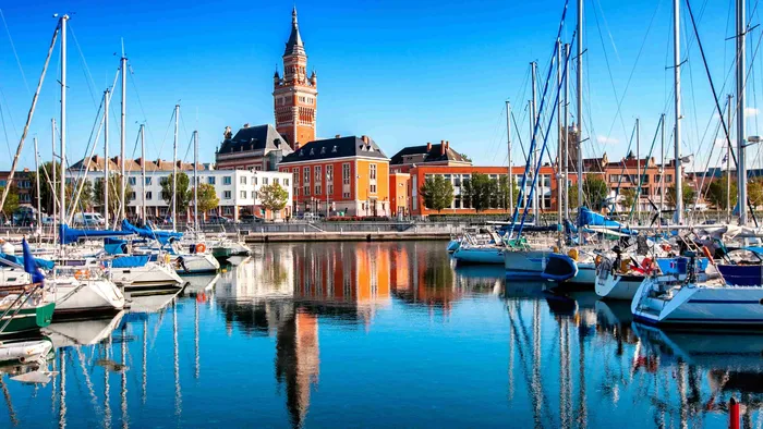 Sailboats docked at a calm harbor reflecting buildings and a tower in Dunkirk at The Originals City Hotel du Parc Gravelines