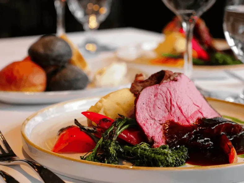 Plate of roast meat, potatoes, vegetables, and sauce, with wine glasses and another plate in background.