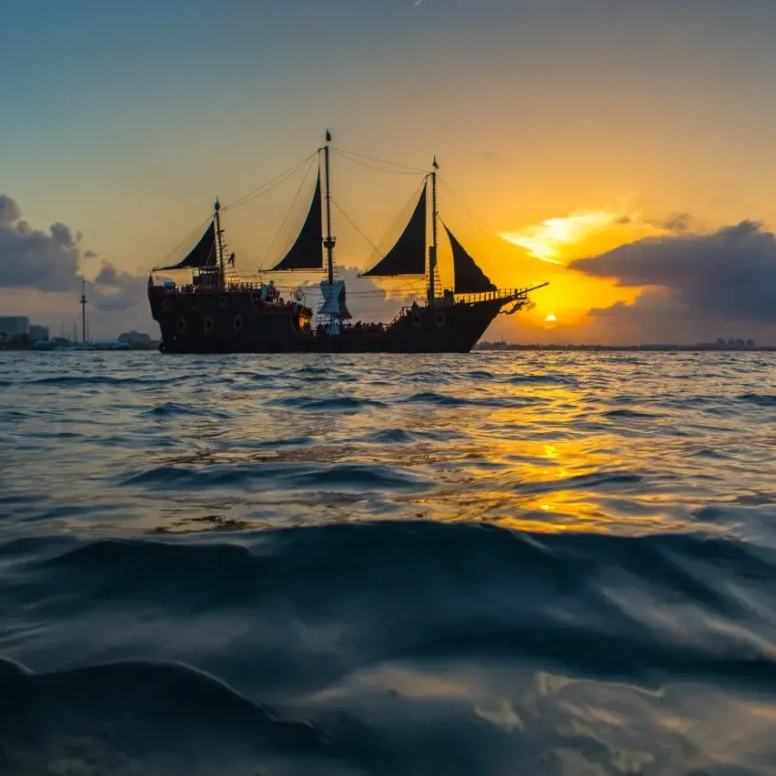 Barco pirata navegando al atardecer con cielo dorado y nubes dramáticas reflejándose en el mar tranquilo