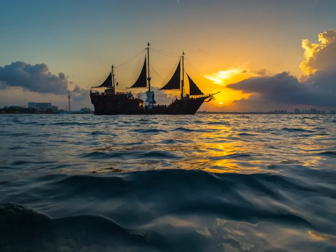 Barco pirata navegando al atardecer con cielo dorado y nubes dramáticas reflejándose en el mar tranquilo