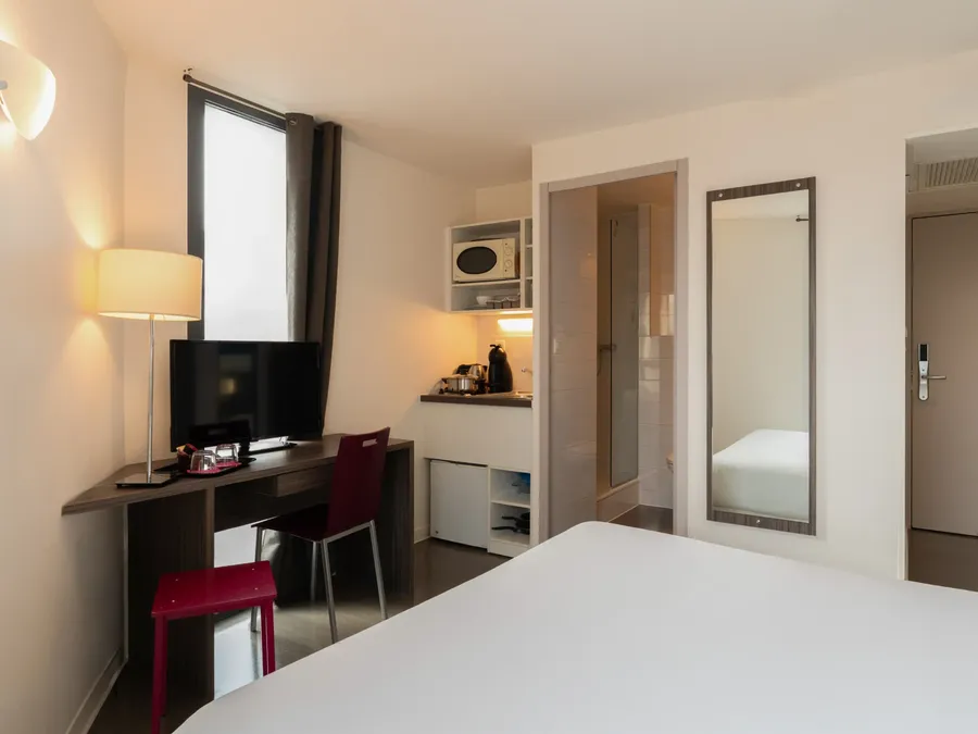 Modern room with desk, chair, TV, microwave, and white bed at Appart’Hôtel Hévéa.