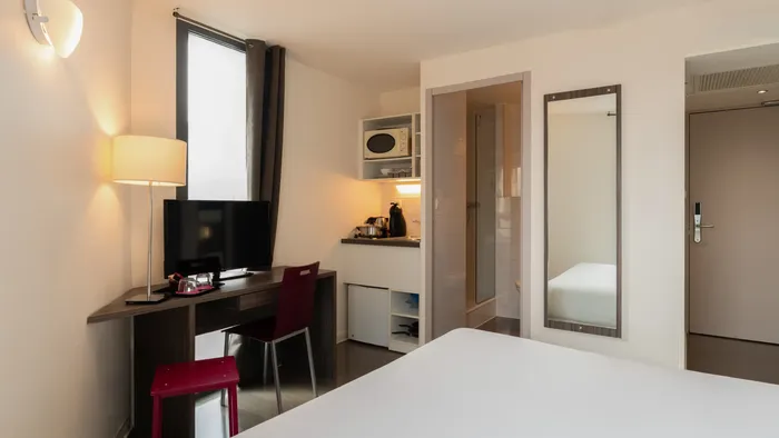 Modern room with desk, chair, TV, microwave, and white bed at Appart’Hôtel Hévéa.