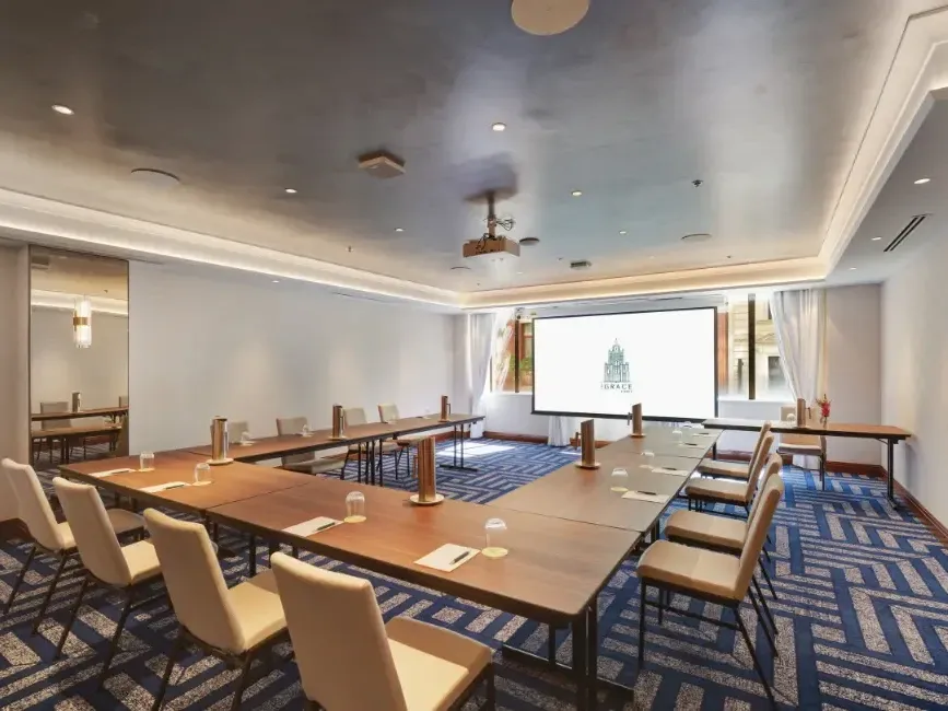 Modern conference room with U-shaped table arrangement, beige chairs, projection screen, and blue patterned carpet.