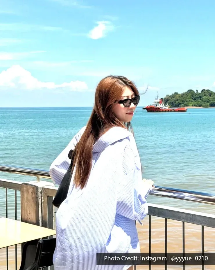 Person posing by waterfront with boat offshore.