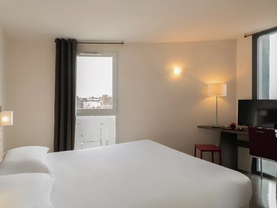 Standard Studio with white bed, desk, chair, and balcony at Appart’Hôtel Hévéa in Valence.