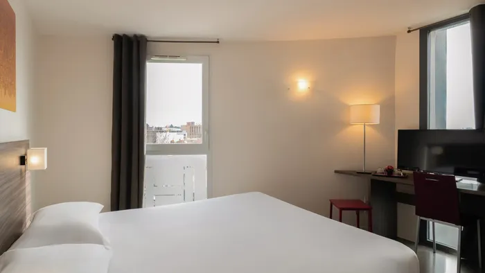White room with bed, desk, chair, and balcony at Appart’Hôtel Hévéa.