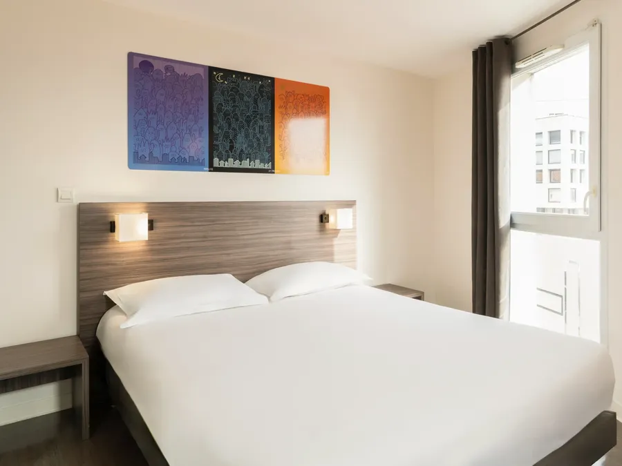 A white bed with two pillows under a wall-mounted lamp in a Standard Studio at Appart’Hôtel Hévéa in Valence.