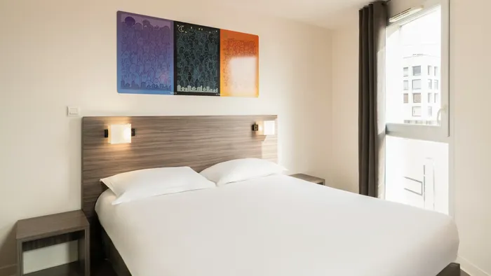 Modern hotel bedroom with bed, nightstands, artwork, and window at Appart’Hôtel Hévéa.