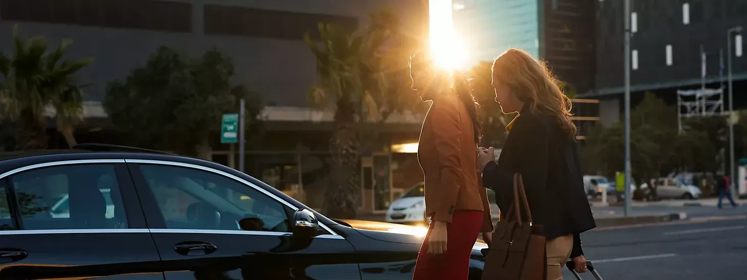 Two women standing next to a black car with sun flare, palm trees, and buildings in the background.