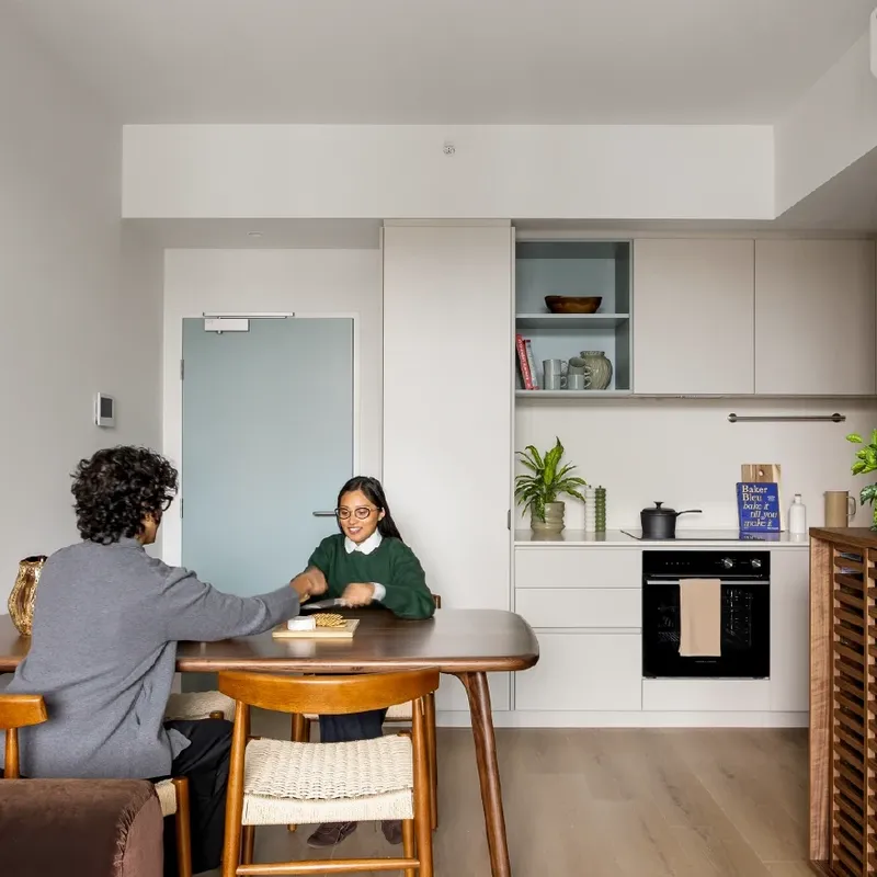 Two students talking at a dining table in a modern kitchen area with wooden furniture and appliances.