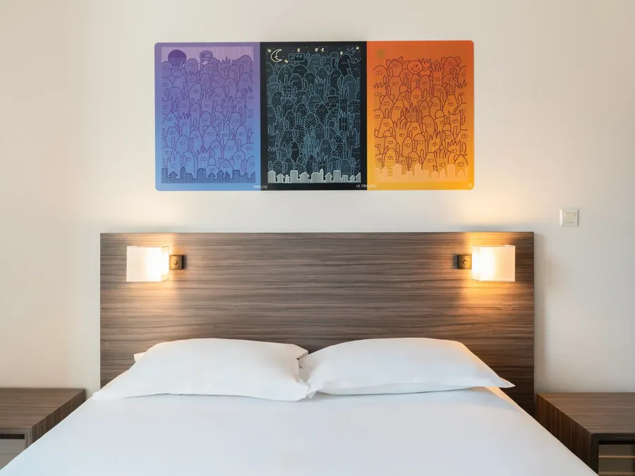 White bed with two pillows and wooden headboard with lit lamps at Appart’Hôtel Hévéa in Valence.