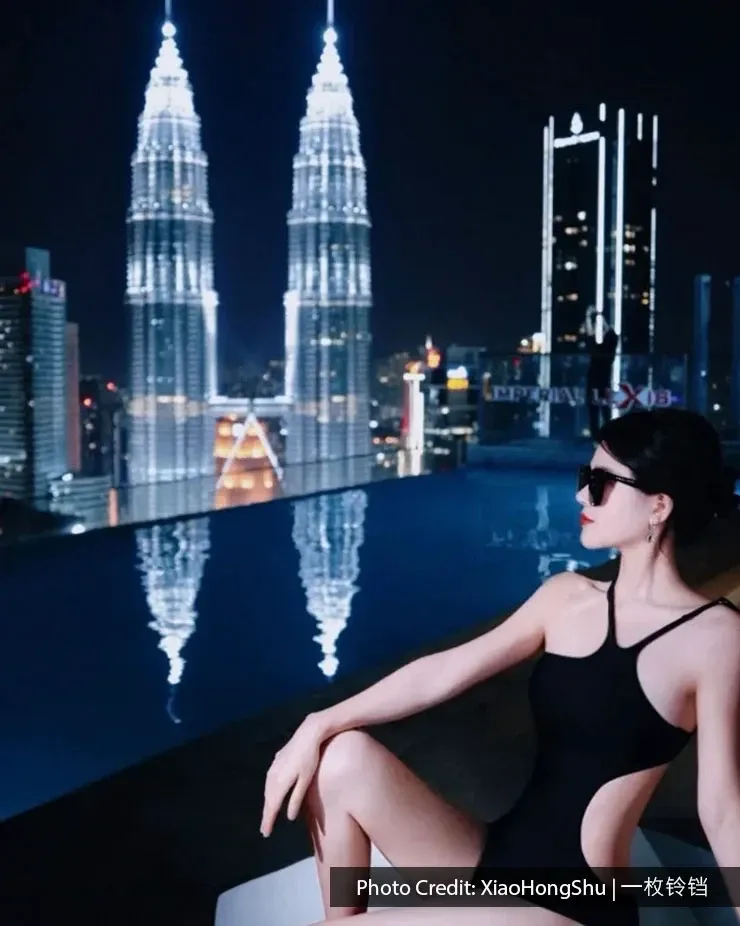 A woman poses by the pool at night, overlooking the brightly lit Petronas Twin Towers.