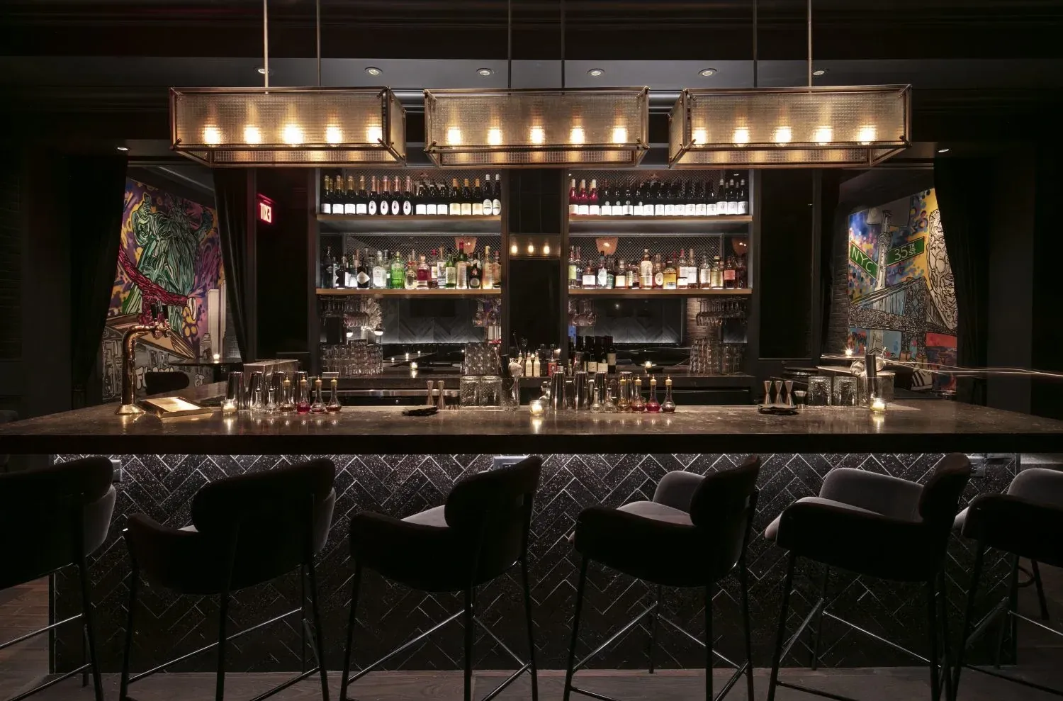 A dimly lit bar with a long counter and stools, shelves of bottles in the background, and artwork on the walls.