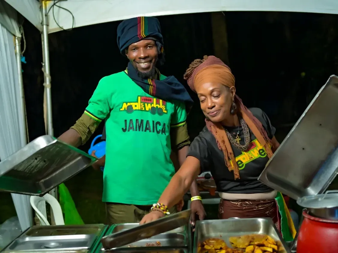 Two food vendors smiling and serving hot meals from chafing dishes at a night event near Jamaica Pegasus Hotel